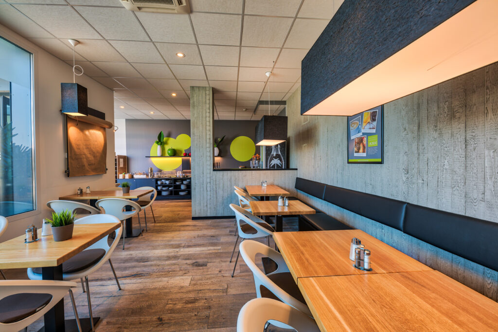 Ein Blick in den hellen, modernen Frühstücksraum in warmen Holz-Tönen. Zu sehen sind verschiedene Sitzmöglichkeiten mit Stühlen und Bänken. Große Fensterscheiben an der linken wand und im Hintergrund ist das Büffet zu erkennen.