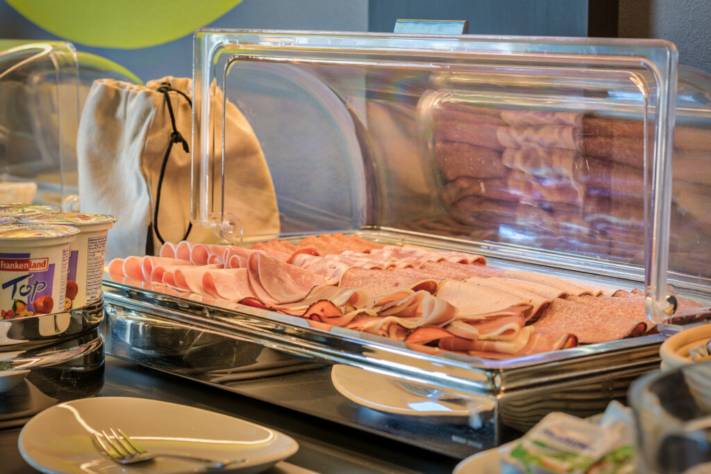 Das Frühstücksbüffet mit einer Nahaufnahme der Wurstplatte, die acht verschiedene Wurst- und Fleischwaren bietet. Am Ran sieht man Joghurtbecher und Butter-Sachets.