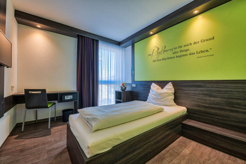 Die Einzelzimmer im EconoMy-Hotel Ulm sind in warmen Brauntönen, weiß und mit hellgrünen Akzenten gehalten. Auf der grünen Rückwand hinter dem Bett steht das Zitat "Der Rhythmus ist für mich der Grund aller Dinge. Mit dem Rhythmus beginnt das Leben." von Herbert von Karajan. Das Zimmer ist mit einem Bodentiefen Fenster mit einem transparenten, sowie einem Vorhang zur Verdunklung ausgestattet. Es gibt einen kleinen Schreibtisch mit Safe, sowie einen Fernseher an der Wand. Rechts und links vom Bett befinden sich Ablagemöglichkeiten.