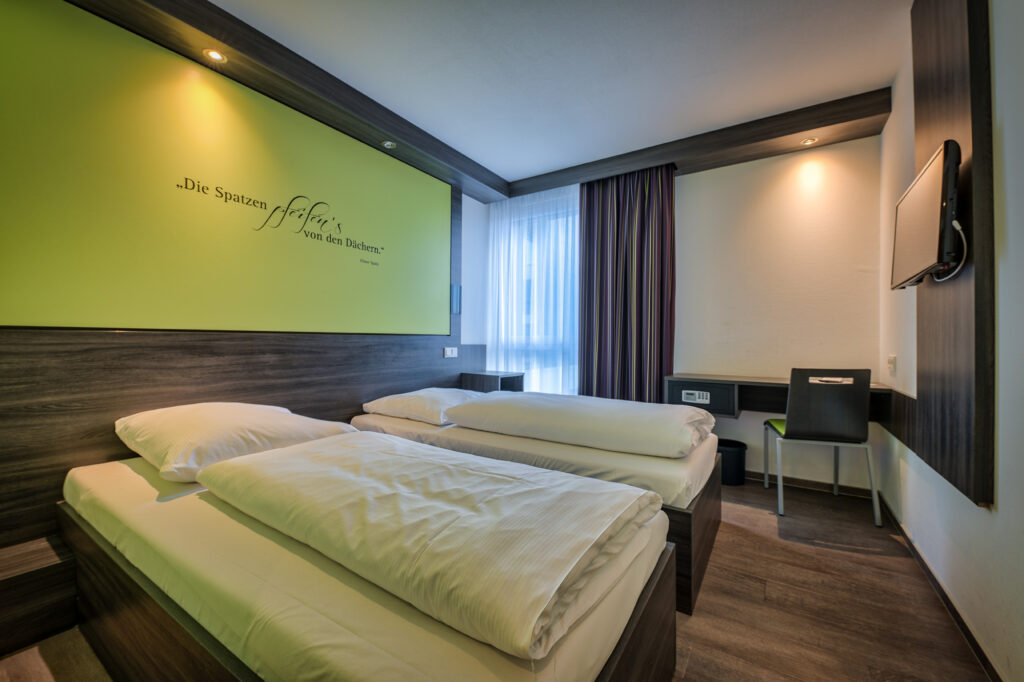 Das Foto zeigt eines der Doppelzimmer im EconoMy-Hotel Ulm. Die Betten stehen hier getrennt, lassen sich aber auch zusammenschieben. Weiterhin befindet sich im Raum ein bodentiefes Fenster mit einem transparenten, sowie einem blickdichten Vorhang. Außerdem ein kleiner Schreibtisch mit Stuhl und Safe, sowie ein Fernseher an der Wand. Der Raum ist in weiß, dunklebraunem Holz und hellem Grün gehalten und an der Wand über den Betten ist der Spruch "Die Spatzen pfeifen's von den Dächern" zu lesen.