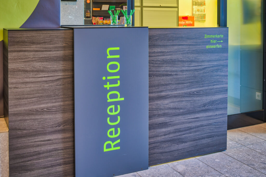 Die Rezeption im Eingangsbereich des EconoMy-Hotels Ulm ist über einen leuchtend grünen Schriftzug auf dem Tresen gekennzeichnet. Auf der rechten Seite des Tresens können Zimmerkarten beim Check-out eingeworfen werden. Der Bereich ist mit einem Schriftzug und Pfeil gekennzeichnet. Auf dem Tresen befinden sich Kugelschreiber. Im Hintergrund ist ein Erste Hilfe Kasten erkennbar.
