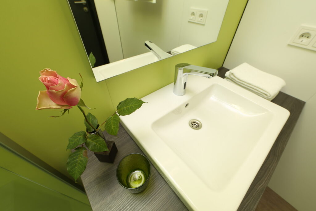A close-up of the washbasin in one of the single rooms. Visible is a light green wall, a modern white sink mounted on a dark brown vanity, fresh white towels, and a vase with a single rose for decoration.