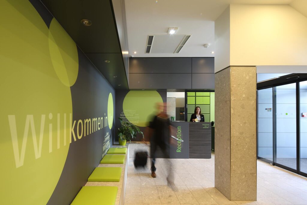 A wide-angle shot of the entire entrance area. On the right, you can see the large glass door leading outside. Straight ahead is the reception desk, staffed by a friendly, smiling employee. Directly opposite the entrance, the word “Welcome” is prominently displayed on the wall, along with some seating options. In the center of the room stands a supporting column. The brightly lit space features light marble, dark grey, and light green tones.