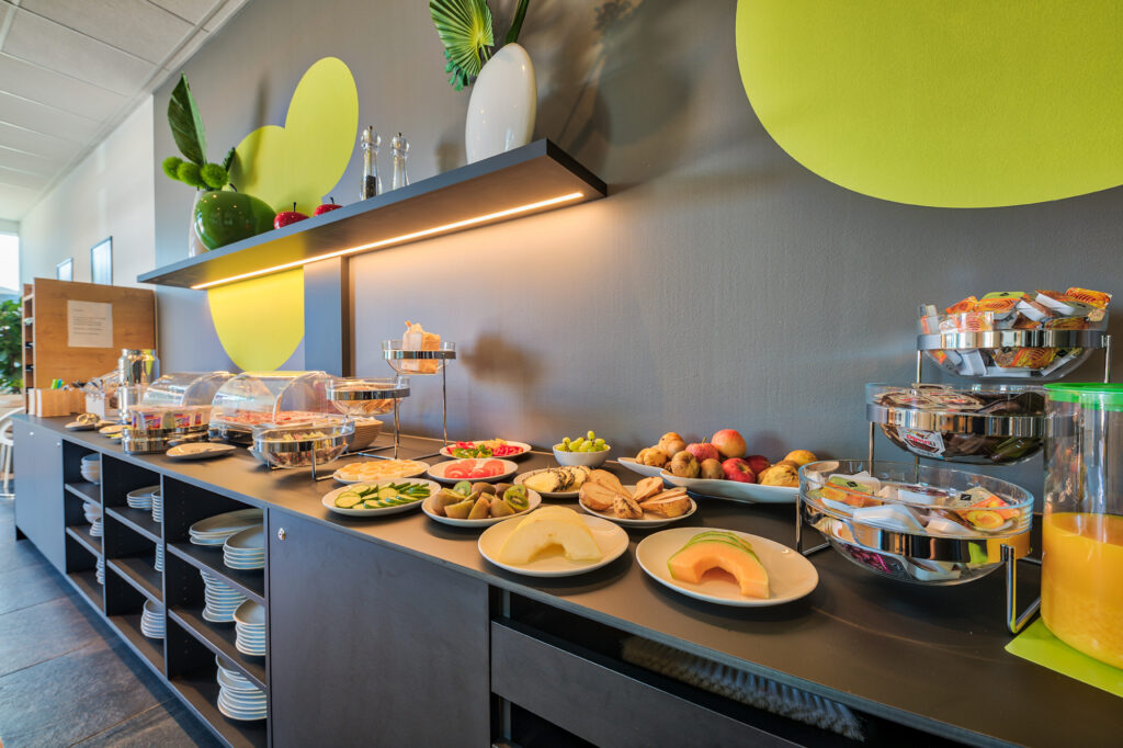 An overview of the breakfast buffet. Visible are fresh fruits such as melon, kiwi, pear, apples, pineapple, and grapes. Fresh vegetables include cucumber, tomato, and bell pepper. Boiled eggs are also displayed, and in the background—slightly out of focus—you can spot a selection of cold cuts and cheeses, yogurts, and more.