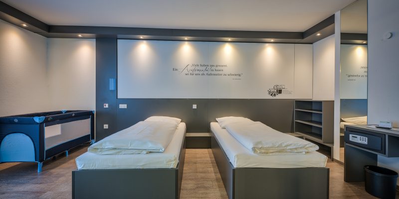 Das Bild zeigt einen hellen, geräumigen Raum mit zwei Einzelbetten un einem Baby-Beistellbett. Der Raum ist in weiß und hellgrau gehalten, mit einem Boden mit Holz-Optik. Neben den Betten sieht man an der Wand einen großen Spiegel und einen Schreibtisch mit Safe.
