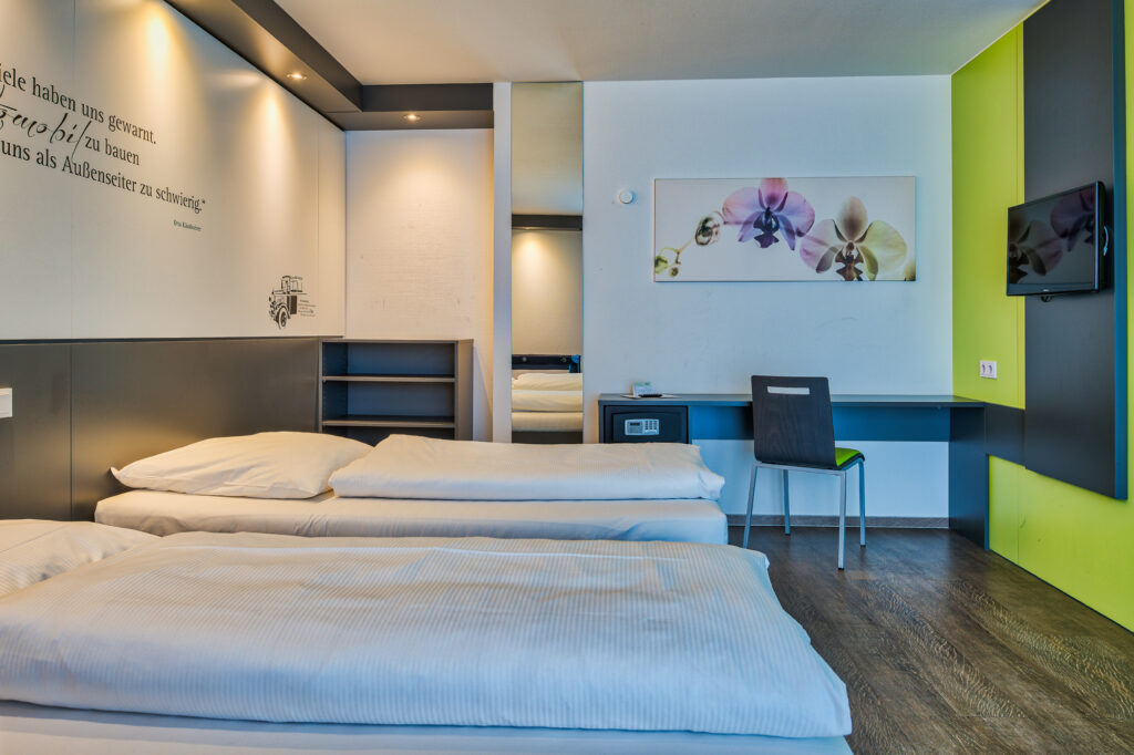 The image shows a bright, spacious room with two single beds. The room is decorated in white and light grey tones, with a wood-look floor. On the wall next to the beds, there’s a large mirror and a desk with a safe and chair. A modern flat-screen TV is mounted on the wall opposite the beds.