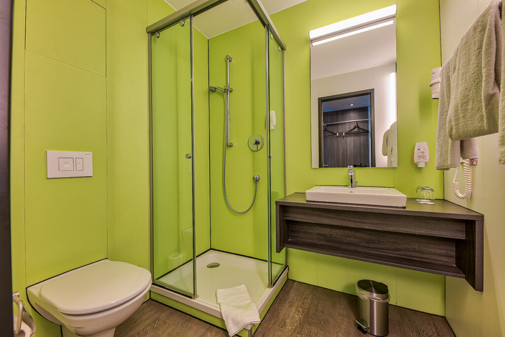 The bathrooms in the double rooms are equipped with a modern toilet, a shower cabin with glass walls and a height-adjustable showerhead, as well as a washbasin with plenty of counter space. You’ll also find a large illuminated mirror, soap, a hairdryer, glasses, and a small waste bin. The bathroom walls are painted in a light green tone.