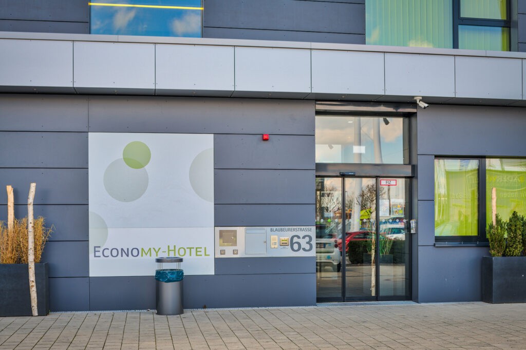 The entrance to the EconoMy Hotel Ulm is at ground level and features a large automatic sliding glass door. The house number is clearly visible, and a large sign identifies the building as the EconoMy Hotel. A trash bin and an ashtray are available in front of the hotel.